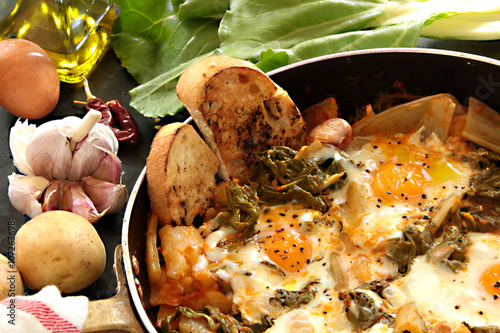 Acquacotta of chard with egges, potatoes and toasted bread. Around: potatoes, egg, chard, garlic, a bottle of olive oil, chilli pepper and a cotton rag.