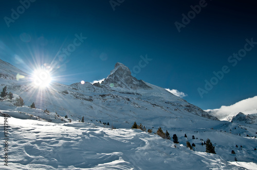 Fotografie View on snowy Matterhorn peak in sunset time with snow and forest trees in foreground and blue sky and sun in background, Switzerland