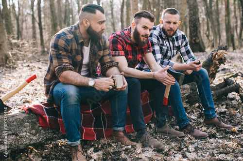 Three bearded men smoke, drink coffee and relax after work in the forest