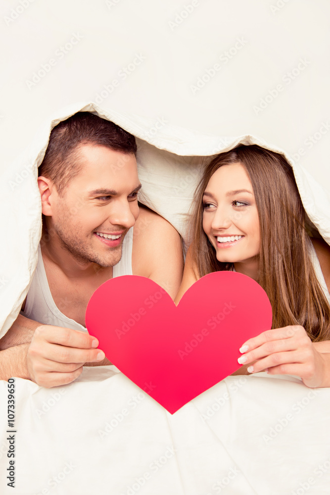Happy couple lying under blanket and holding bid paper heart