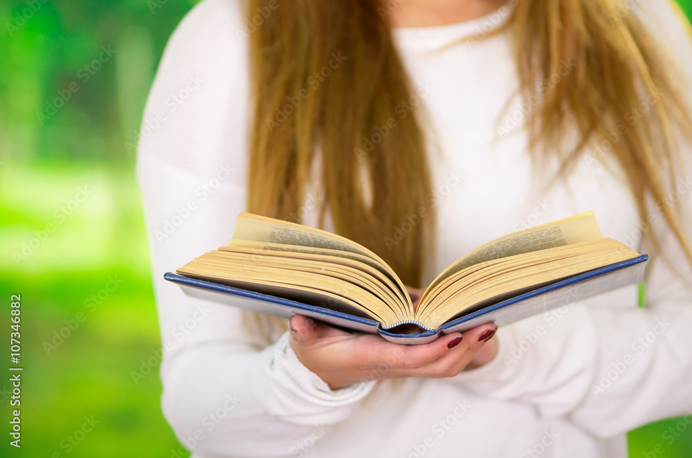 Closeup open book held by woman wearing white sweater and long hair visible, garden background Closeup open book held by woman wearing white sweater and long hair visible, garden background
