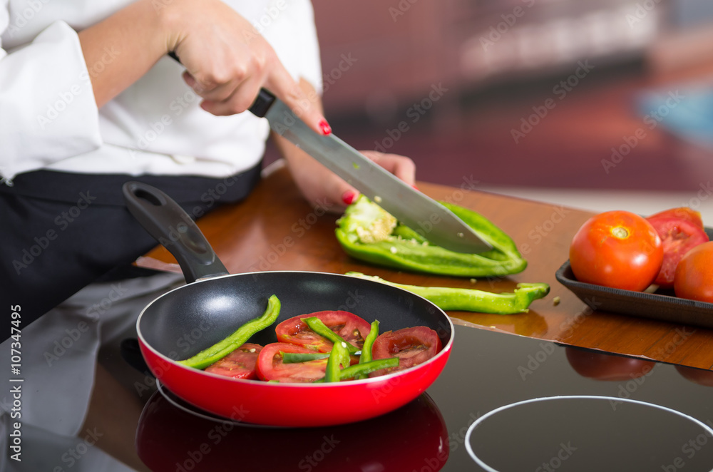 Closeup chef cutting green peppers on wooden surface with red skillet in front frying up vegetables