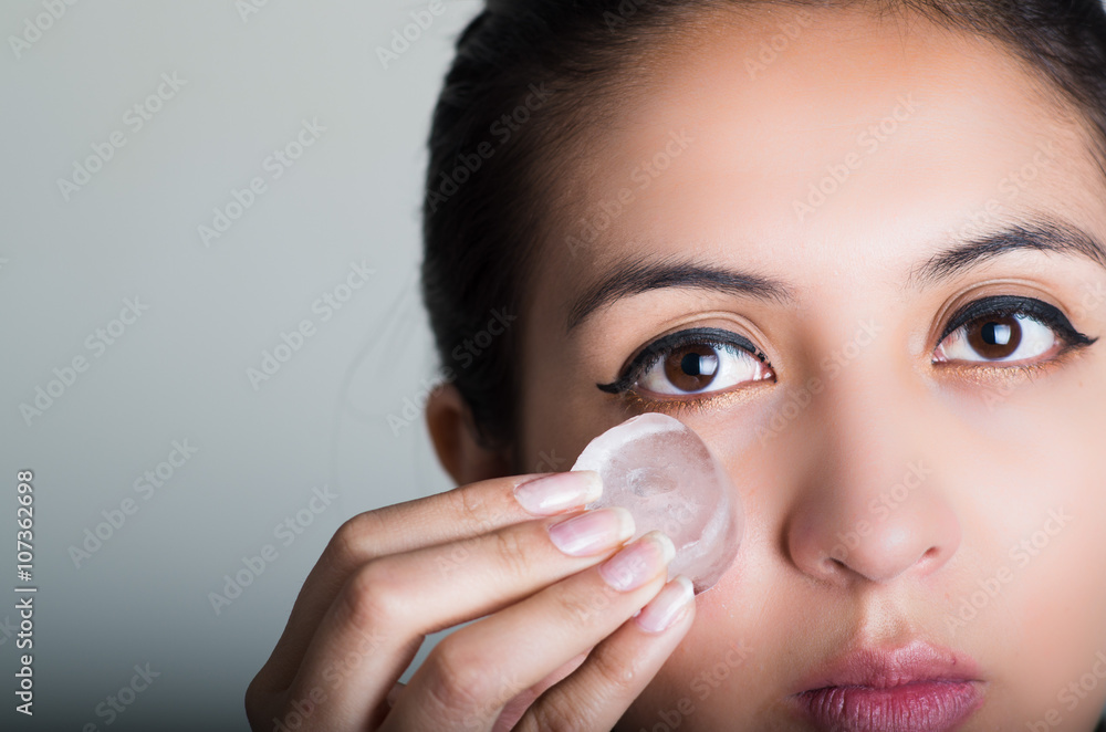 Gorgeous brunette model rubbing ice cube against cheek underneath eye, grey background Gorgeous brunette model rubbing ice cube against cheek underneath eye, grey background
