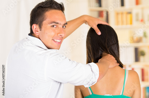 Male physio therapist with woman client sitting seen from behind, helping patient applying pressure massaging neck and back of head, blurry clinic background