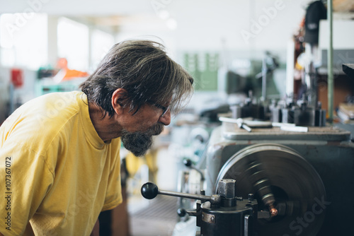 Portrait of a serious bearded worker with glasses doing some job on metalworking machine. Selective focus.
