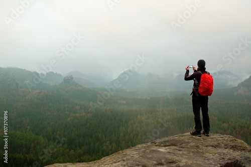 Tourist with backpack takes photos with smart phone of rainy vally. Dreamy foggy valley below