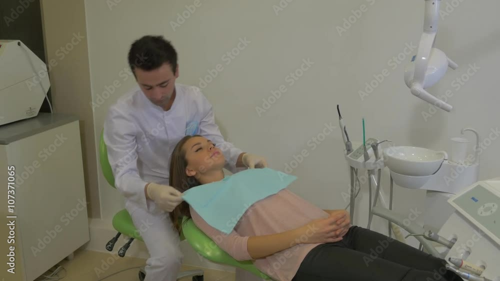 Male Dentist Fixing a Paper Napkin on Chest Put the Mask on and Starts to Examine a Teeth Client Young Woman is Sitting in a Dentist Chair Dental Clinic Male Dentist Fixing a Paper Napkin on Chest Put the Mask on and Starts to Examine a Teeth Client Young Woman is Sitting in a Dentist Chair Dental Clinic