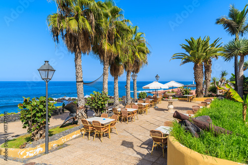 Palm trees and restaurant tables on coastal promenade in Puerto de la Cruz town, Tenerife, Canary Islands, Spain