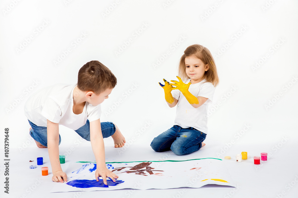Two pretty child friends boy and girl in white shirts and blue jeans,  trendy hair style, barefoot, drawing pictures on white sheet of paper by  paints isolated on white. Studio shot. Stock
