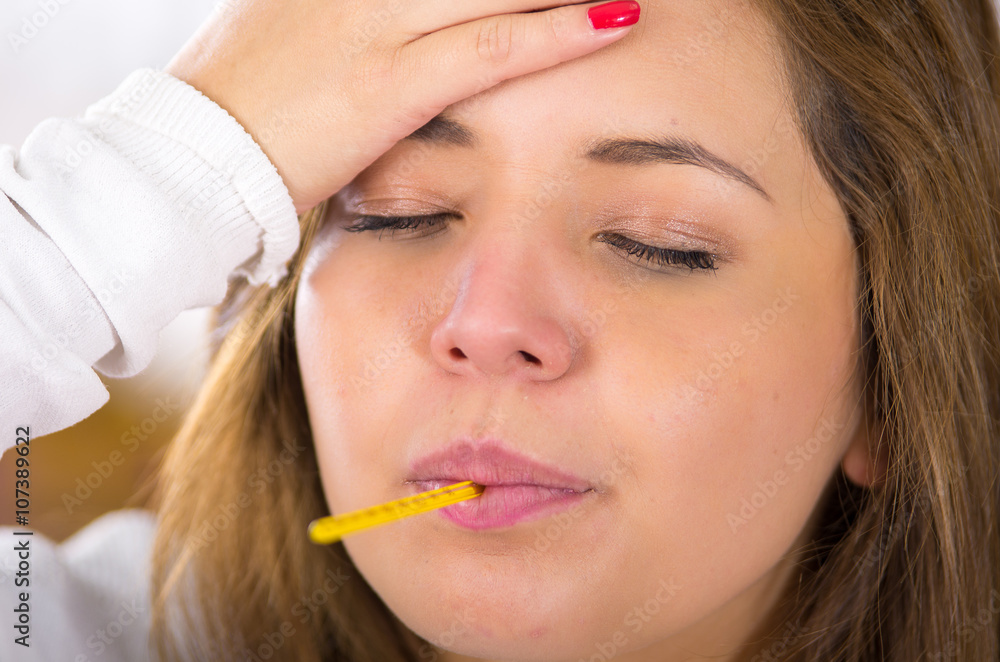 Closeup headshot brunette sick with the flu holding her head, thermometer in mouth Closeup headshot brunette sick with the flu holding her head, thermometer in mouth