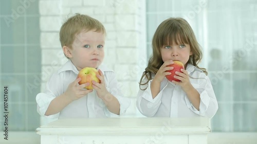 Little boy and girl eat red apples.We look at each other and then leave. Children in white shirts next to the table in a bright room. Against the background of light brick wall and the window.