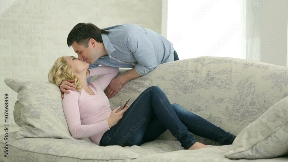 A happily married couple. The husband and wife on the couch, talking, kissing, joking, having a good time. Bright room, white brick wall, a chest of drawers. European couple. A happily married couple. The husband and wife on the couch, talking, kissing, joking, having a good time. Bright room, white brick wall, a chest of drawers. European couple.
