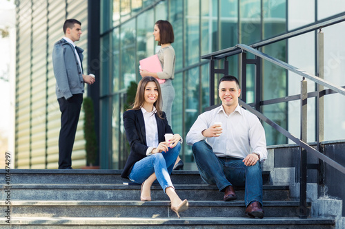 Young businesswoman and businessman sitting on the steps of the office building, drinking coffee and talking. They are looking at camera and smiling. People in the background © bokan