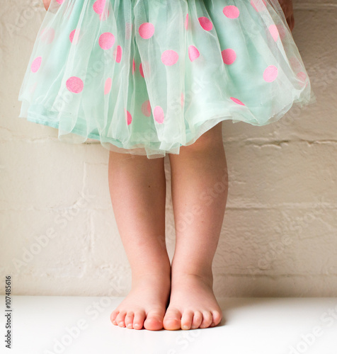Toddler in green and pink polka dot skirt against white brick wall (cropped)
