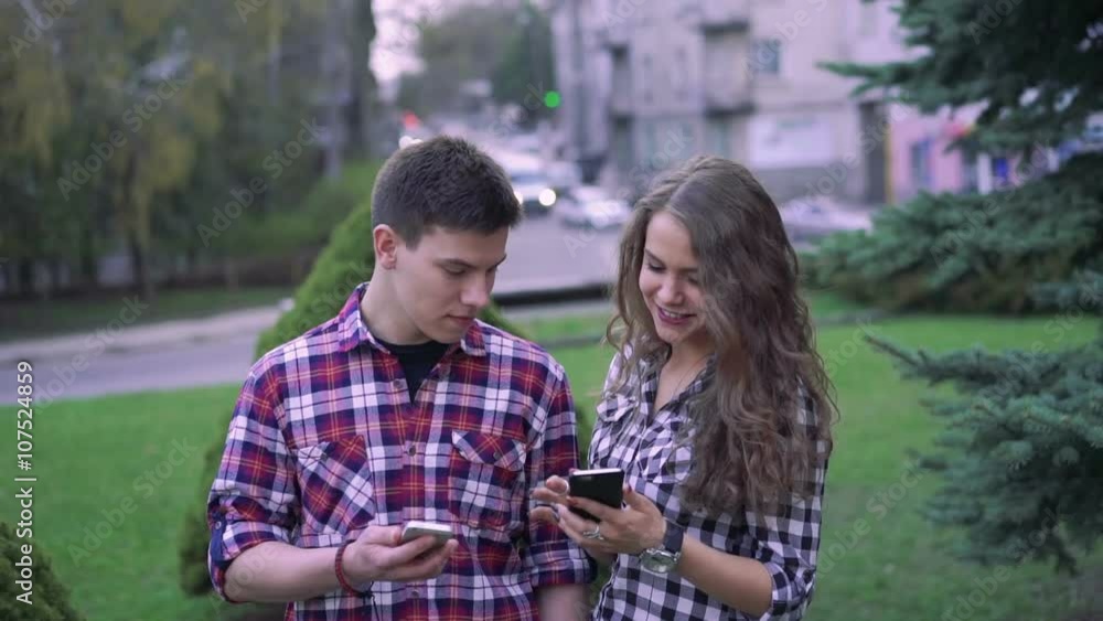 Happy couple looking on phones, laughing near trees in the center Happy couple looking on phones, laughing near trees in the center