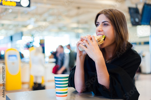 Young woman at international airport, drinking coffee and eating a sandwich while waiting for her flight. Female passenger at terminal, indoors.