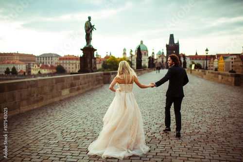 Happy cheerful newlywed couple running on bridge in Prague, hone