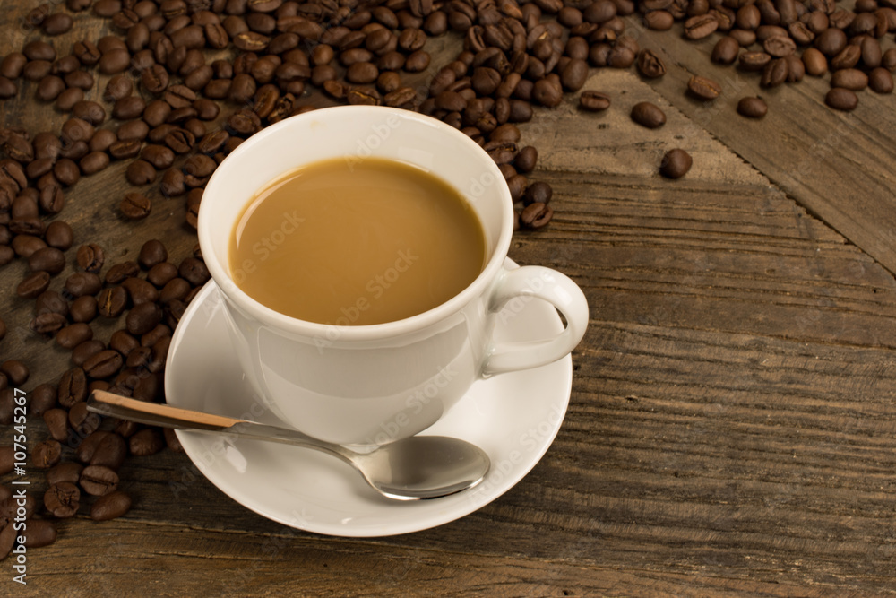 Coffee with milk in white cup with silver spoon and fresh coffee beans on rustic wooden table. Coffee with milk in white cup with silver spoon and fresh coffee beans on rustic wooden table.
