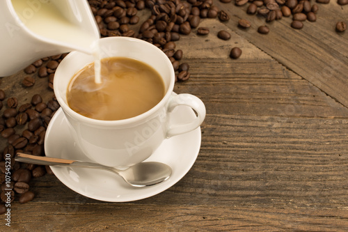 Pouring milk in a cup of coffee and beans on wooden background