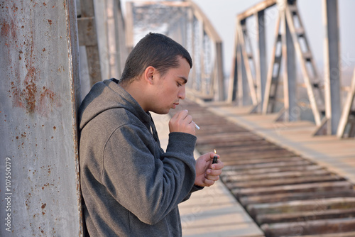 Young man wearing a hoodie standing by a bridge and lighting a c