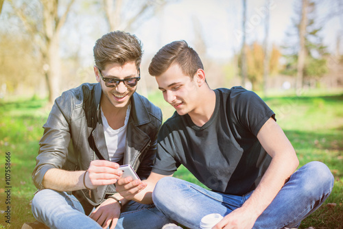 Smiling friends with smartphones sitting on grass in park