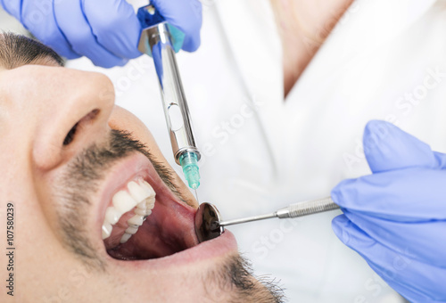 Dentist holding a syringe and angled mirror, anesthetizing his patient at dental clinic. Dental care concept.