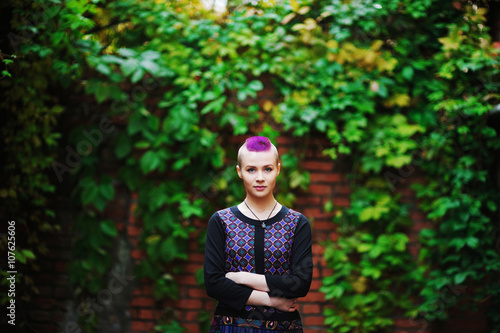 Beautiful punk girl with a purple mohawk standing against a background of brick wall overgrown with leaves. photo