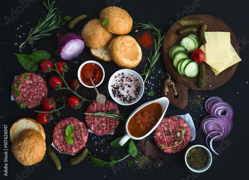 Ingredients for cooking burgers. Raw ground beef meat cutlets  buns  red onion  cherry tomatoes  greens  pickles  tomato sauce  cheese  herbs and spices over black background  top view.