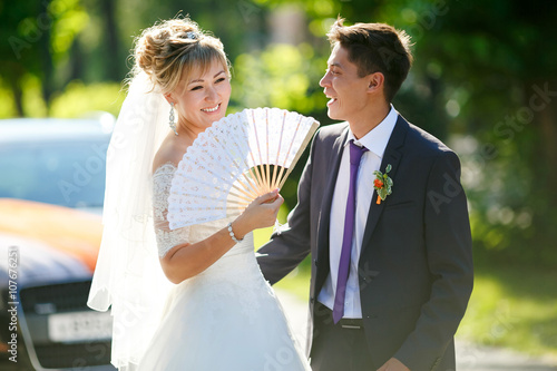 Groom and bride with fan on street illuminated by sun, car in background.