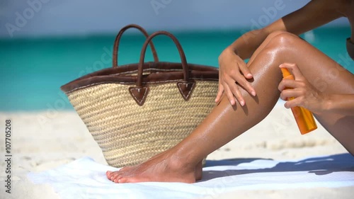 Sunscreen suntan lotion in spray bottle. Young woman in spraying tanning oil on her leg from bottle. Lady is massaging sunscreen lotion while sunbathing at beach. Female model during summer vacation.