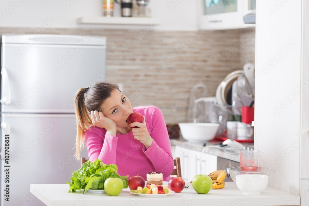 Healthy young woman eating an apple and sitting in kitchen. She is surrounded by fruit. She is not happy about it.