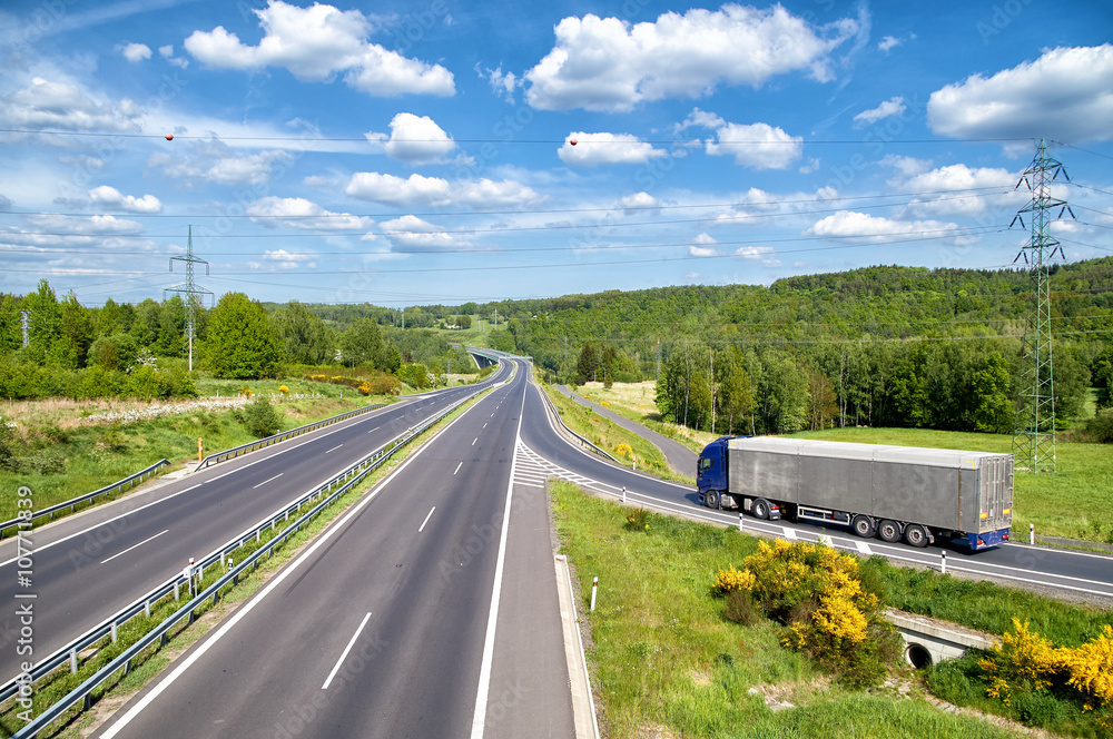 Truck entering on an empty asphalt highway in the landscape. High power line pylons. Electronic toll gate in the distance. Sunny summer day with blue skies and white clouds. View from above.