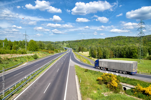 Obraz na płótnie Truck entering on an empty asphalt highway in the landscape. High power line pylons. Electronic toll gate in the distance. Sunny summer day with blue skies and white clouds. View from above. w salonie