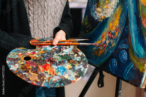 Closeup of paintbrushes and stained mixing  palette in woman hands. Unrecognizable female artist in a workshop holding a stained palette and brush. Closeup of painting tools