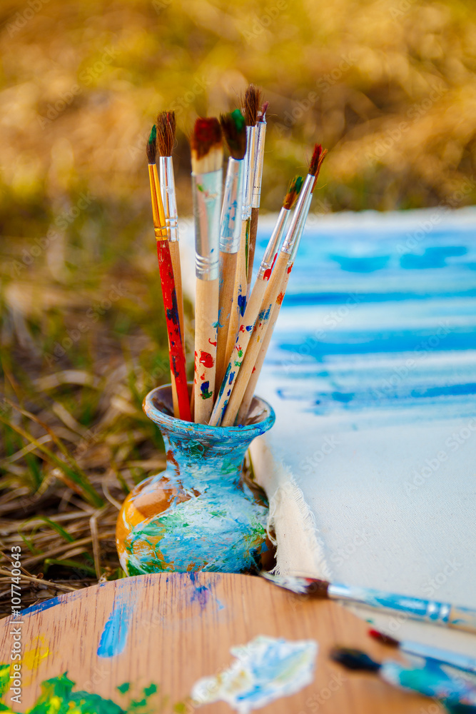 Fototapeta premium Used brushes in the vase near picture with an artist's palette of colorful oil paint for drawing and painting lying on the grass