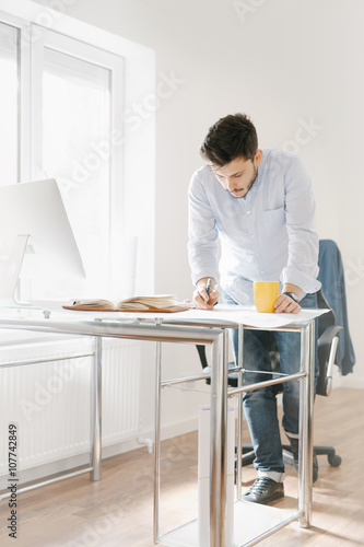 Young man in a modern office design sketching construction project design in a note book computer and drinks coffee