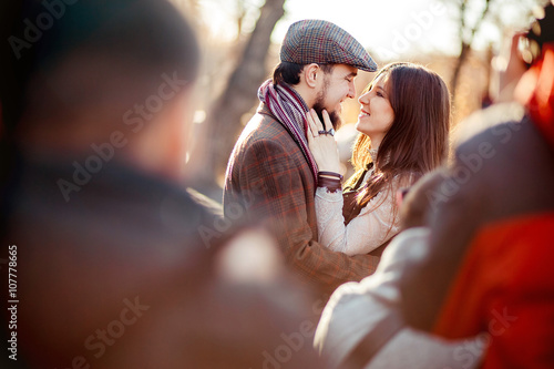 Stylish old fashioned couple among crowd looking at each other outdoors in backlight. Man wearing tweed flat cap, brown clothes.