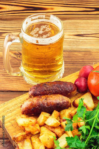 Grilled meat sausages with fried potatoes, fresh produce and mug of beer on wooden board