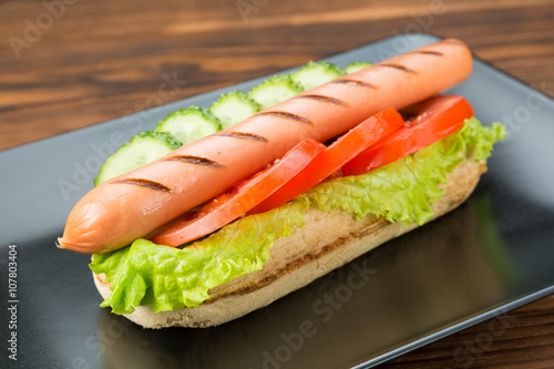 Spicy hotdog with fresh vegetables lying on black rectangular plate