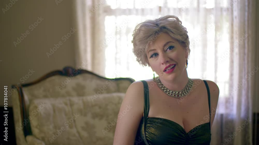 Attractive woman wearing a classic fifties dress and sitting on a chaise lounge looks at camera and smiles. Medium shot, recorded in 4K. Attractive woman wearing a classic fifties dress and sitting on a chaise lounge looks at camera and smiles. Medium shot, recorded in 4K.