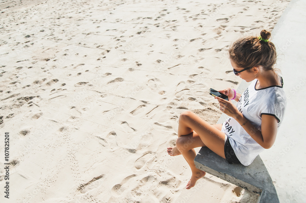 Woman typing text message on smart phone on the beach. Young woman sitting at the beach and chatting in mobile phone.