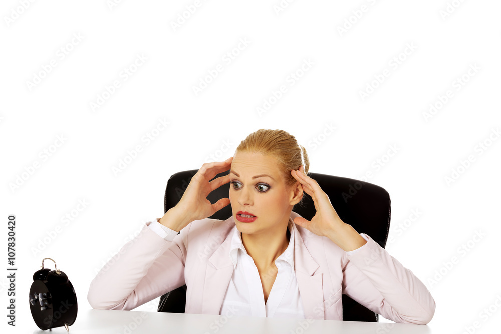 Surprised business woman looking at alarm clock behind the desk Surprised business woman looking at alarm clock behind the desk