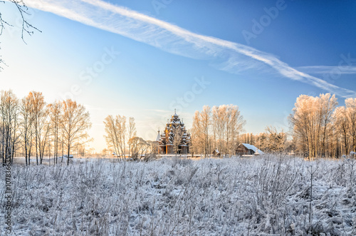A scenery with grass covered with frost and a faraway wooden church behind birches at a very frosty winter day