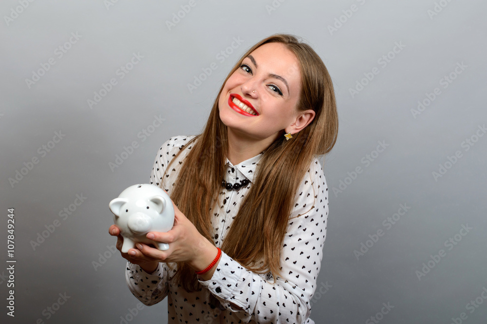 Happy Young Woman Holding Coin And Piggy bank against a grey background Happy Young Woman Holding Coin And Piggy bank against a grey background