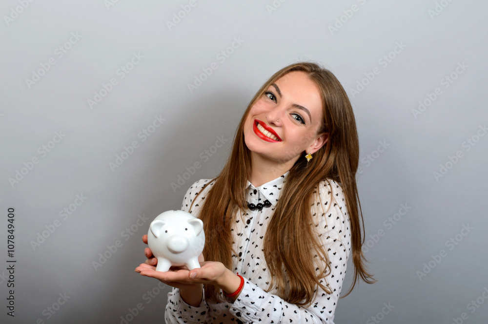 Happy Young Woman Holding Coin And Piggy bank against a grey background Happy Young Woman Holding Coin And Piggy bank against a grey background