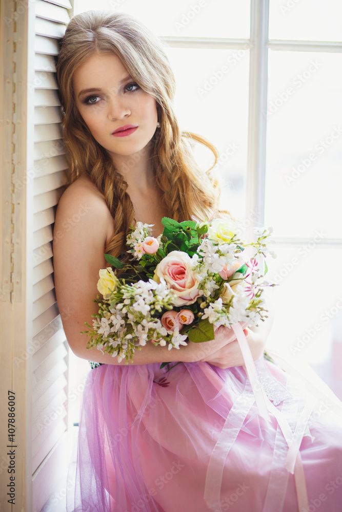 Portrait of pretty young girl in a sweet dress and long blonde hair sitting on a window with flowers composition