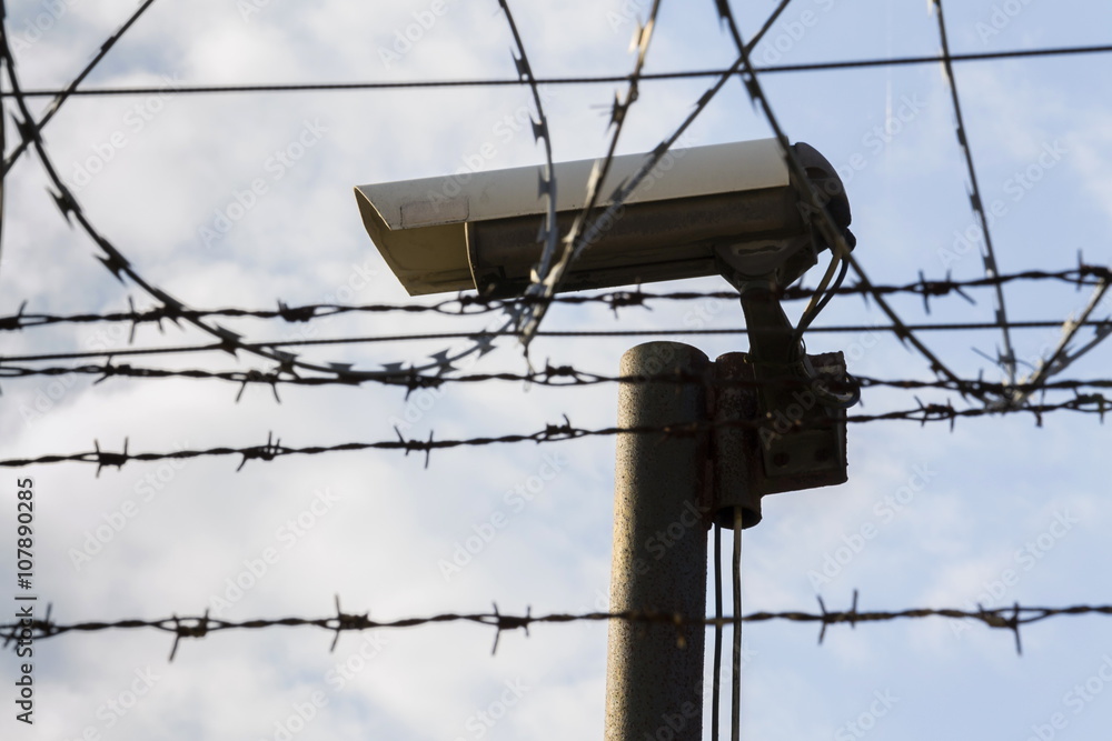 Obraz premium Security camera behind barbed wire fence stretched around prison walls