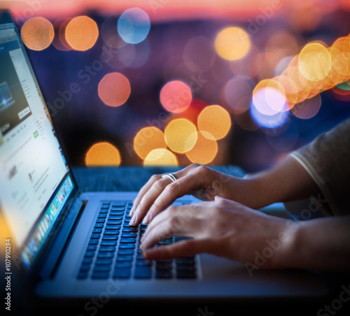 Woman hands typing on laptop keyboard on abstract blurred bokeh of city night light background. Focus in the foreground.