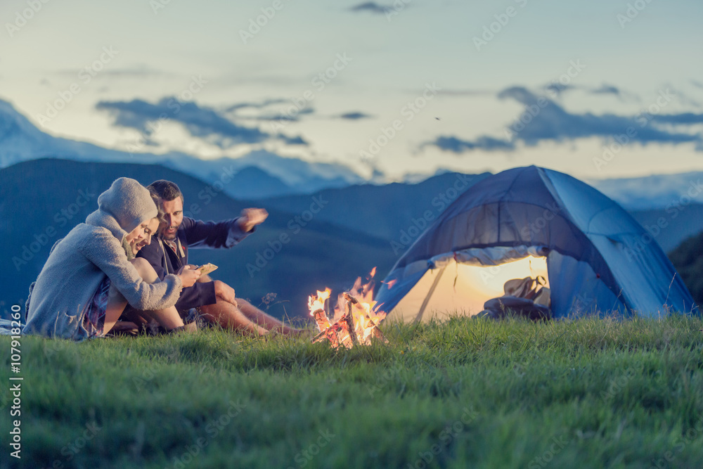 Three friends camping with fire on mountain at sunset - obrazy, fototapety, plakaty 