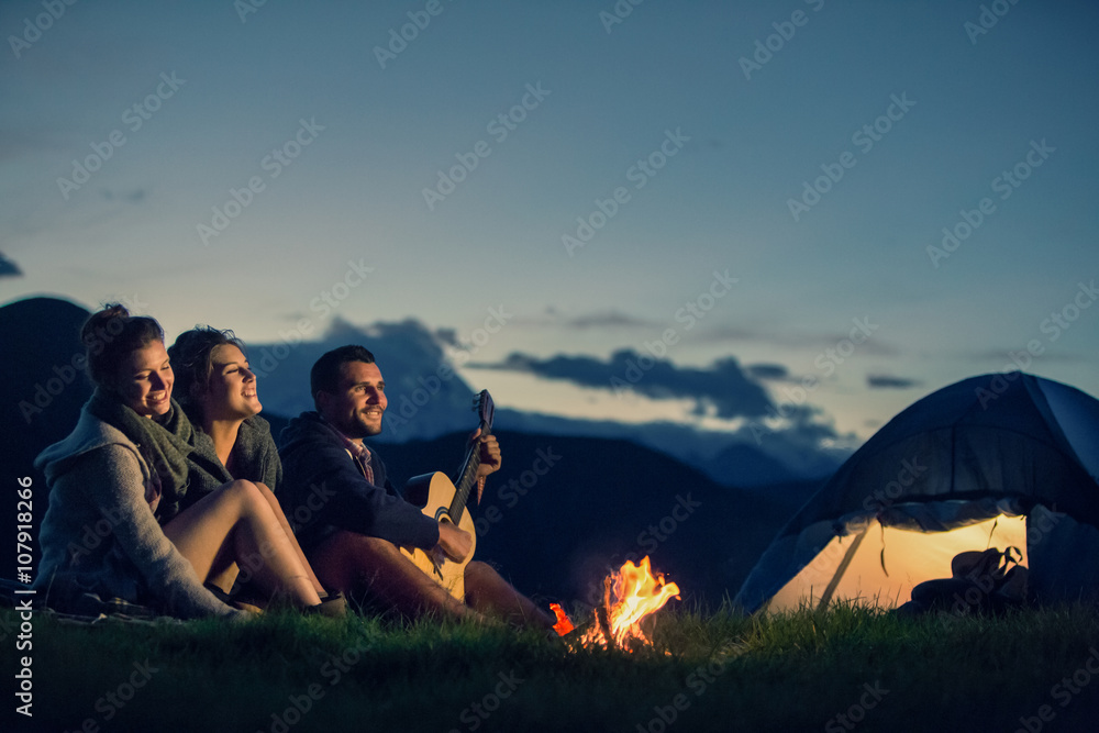 Three friends camping with fire on mountain at sunset - obrazy, fototapety, plakaty 
